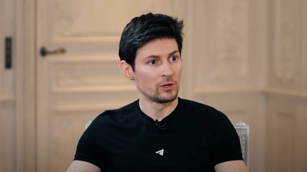 Durov with short dark hair and light skin wearing a black t-shirt with a small white logo, sitting indoors against a beige paneled wall with a decorative door handle visible.