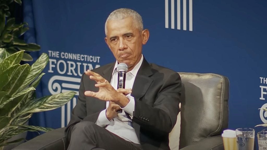 Obama in a black suit holding a microphone sitting on a gray armchair during an event at The Connecticut Forum with plants in the foreground and cups on a table to the side.