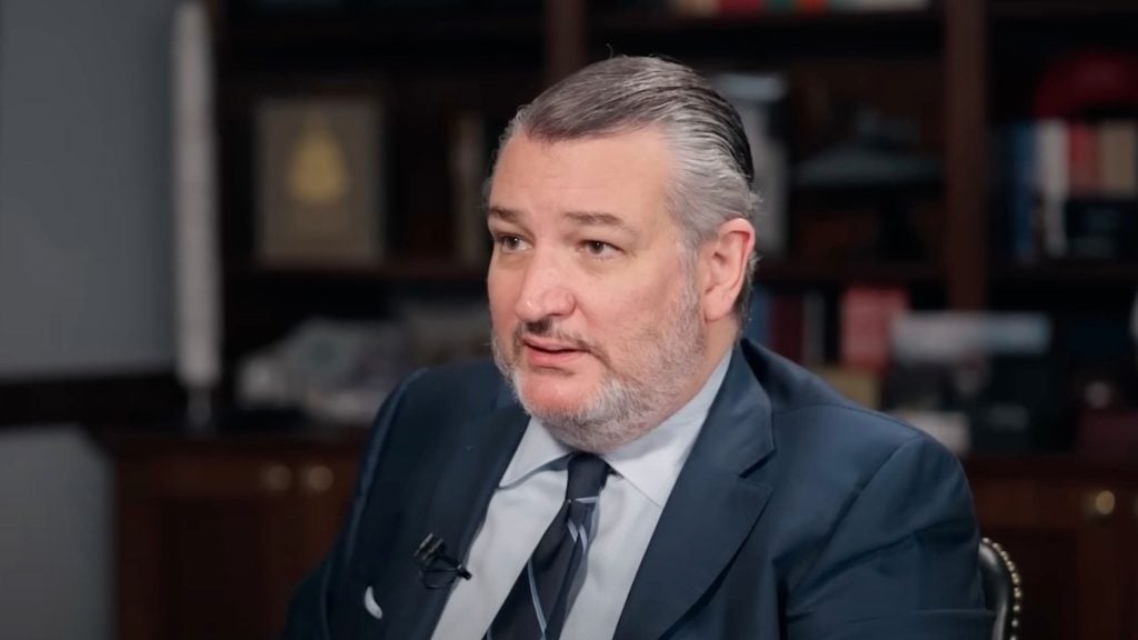 Senator Ted Cruz with gray hair slicked back and a trimmed beard, wearing a navy suit, light blue shirt, and striped tie, sitting in an office with wooden shelves and books in the background.