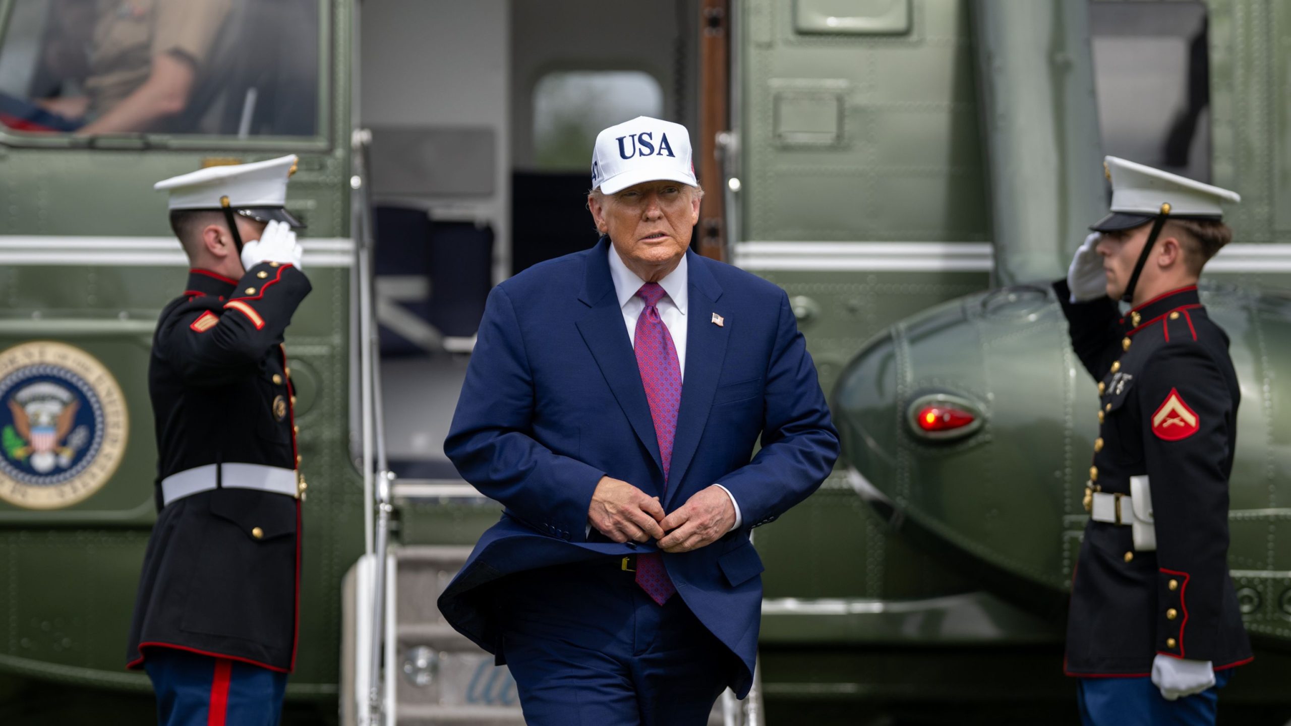 President Trump in a blue suit and red tie wearing a white hat with "USA" printed on it, buttoning his jacket while walking down the stairs of a green helicopter, flanked on both sides by two uniformed Marines saluting.