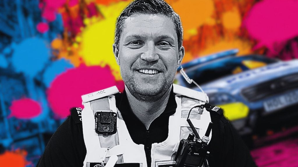 Cooke wearing a white tactical vest with communication equipment, set against a colorful background featuring splashes of pink, yellow, blue, and orange, and a blurred police car with flashing lights.