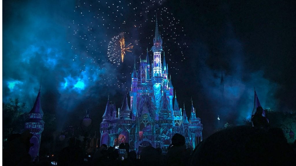 A brightly illuminated castle at night with blue and purple lights and fireworks exploding in the dark sky behind it, surrounded by silhouettes of people watching the display.