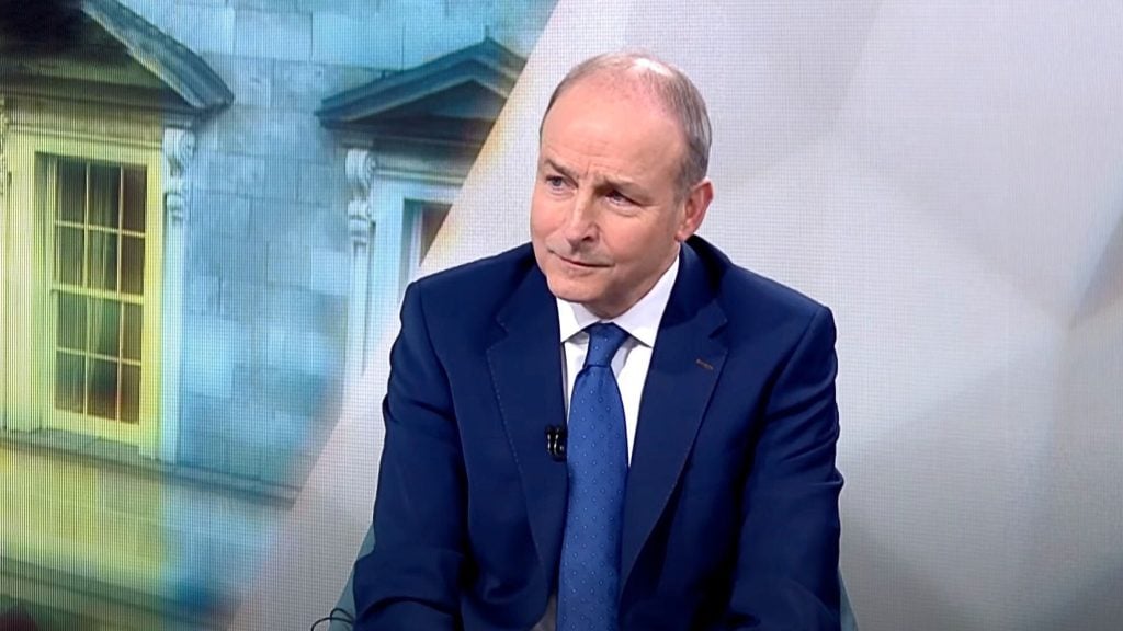 Micheál Martin in a dark blue suit and blue tie sitting and looking slightly to the side against a blurred backdrop of a windowed building facade and a light wall.