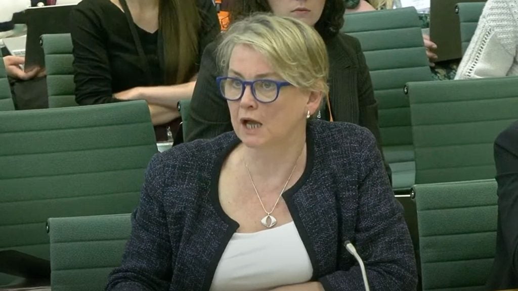 Cooper with blonde hair and blue glasses speaking in a formal setting, seated on a green chair, wearing a dark textured jacket over a white top and a silver necklace, with other people seated behind her.