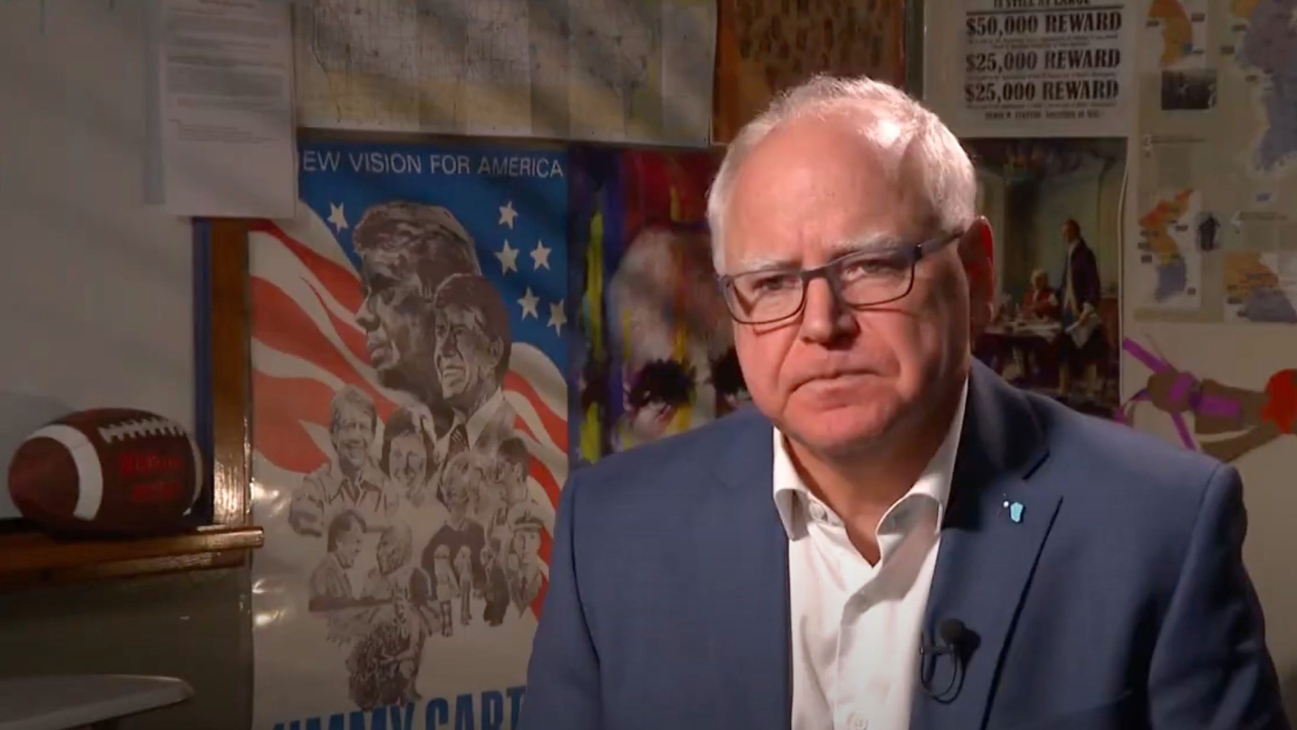 Man with glasses wearing a blue blazer and white shirt, sitting indoors with a microphone clipped to his lapel, various posters and maps visible on the wall behind him including an American-themed poster and a football on a shelf.