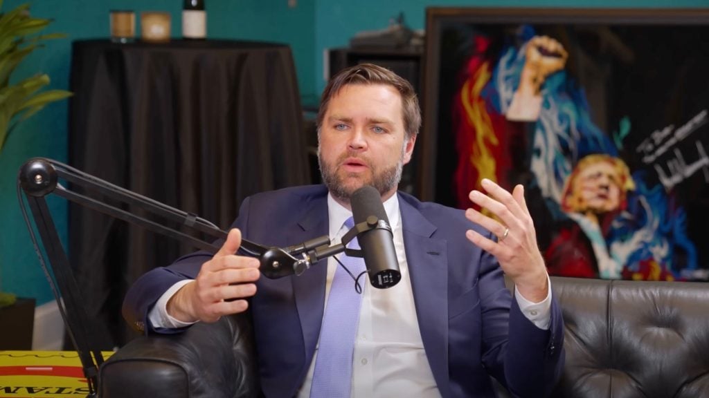 JD Vance in a suit and light blue tie speaking into a microphone on a desk-mounted boom arm while seated on a black leather couch, with colorful artwork and a black-covered object in the background.
