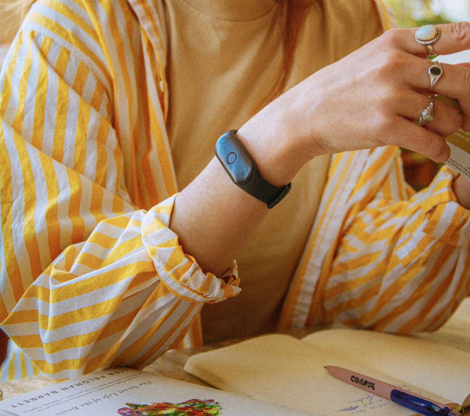 Person wearing a yellow and white striped shirt and a yellow top, with a black fitness tracker on their wrist and multiple rings on their fingers, holding a card or small booklet above an open notebook with a pen.