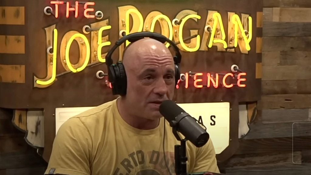 Joe Rogan wearing black headphones and a yellow t-shirt speaking into a microphone in front of a neon sign that reads 'The Joe Rogan Experience' on a wooden wall background.