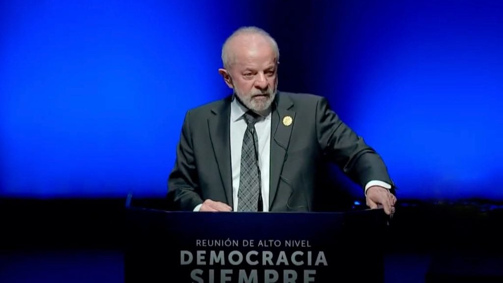 Lula with a gray beard in a dark suit and patterned tie standing at a podium against a deep blue background, with text on the podium reading 'Reunión de alto nivel Democracia Siempre'