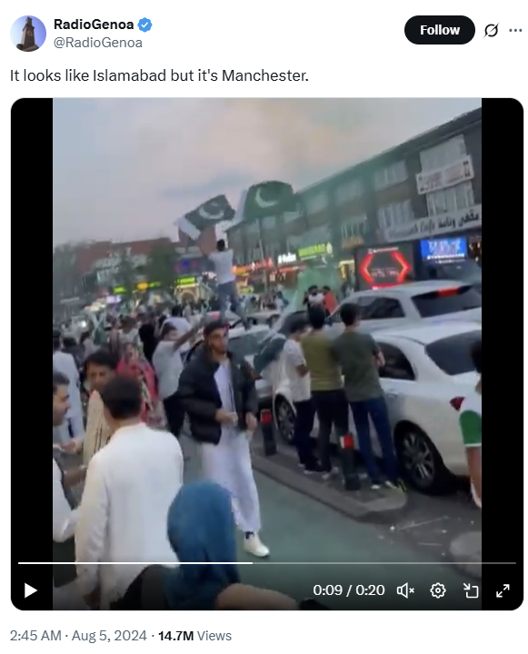 Crowd of people gathered on a busy street with several holding and waving Pakistani flags, surrounded by cars and shops with illuminated signs under a cloudy sky.