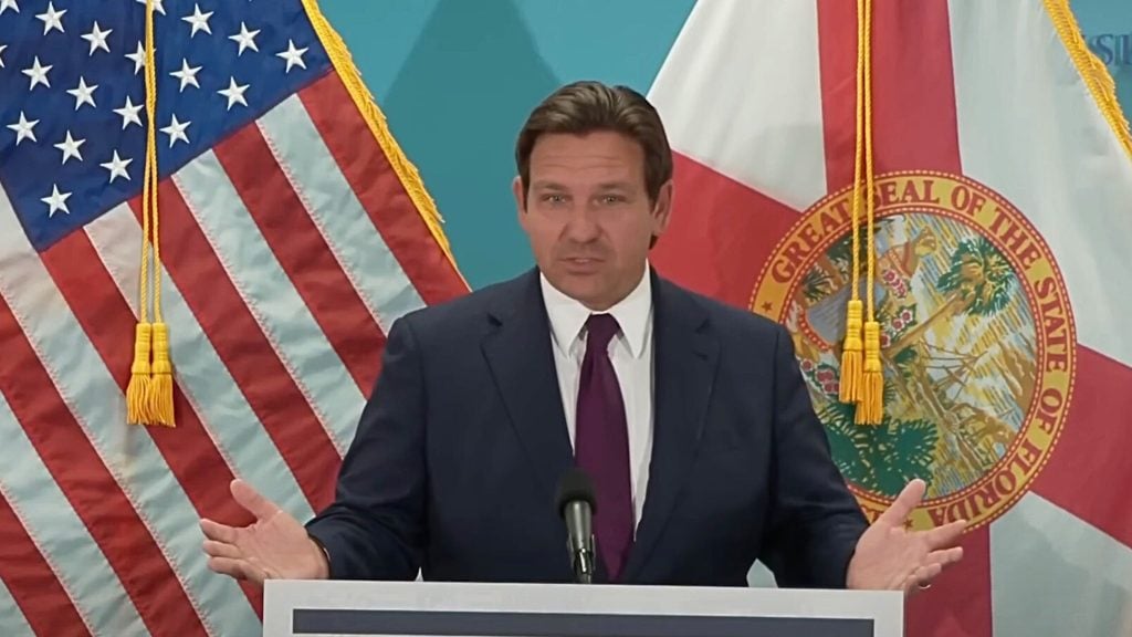 A man in a dark suit and purple tie speaking at a podium with the American flag and the Florida state flag in the background.