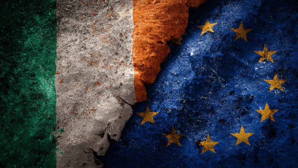 The flags of Ireland and the European Union painted on a cracked, textured concrete surface, with the Irish flag on the left and the EU flag with yellow stars on a blue background on the right.
