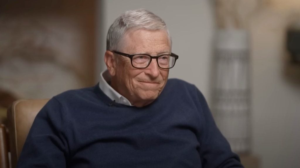 Bill Gates with gray hair and glasses wearing a navy blue sweater over a white collared shirt, sitting and smiling slightly indoors.