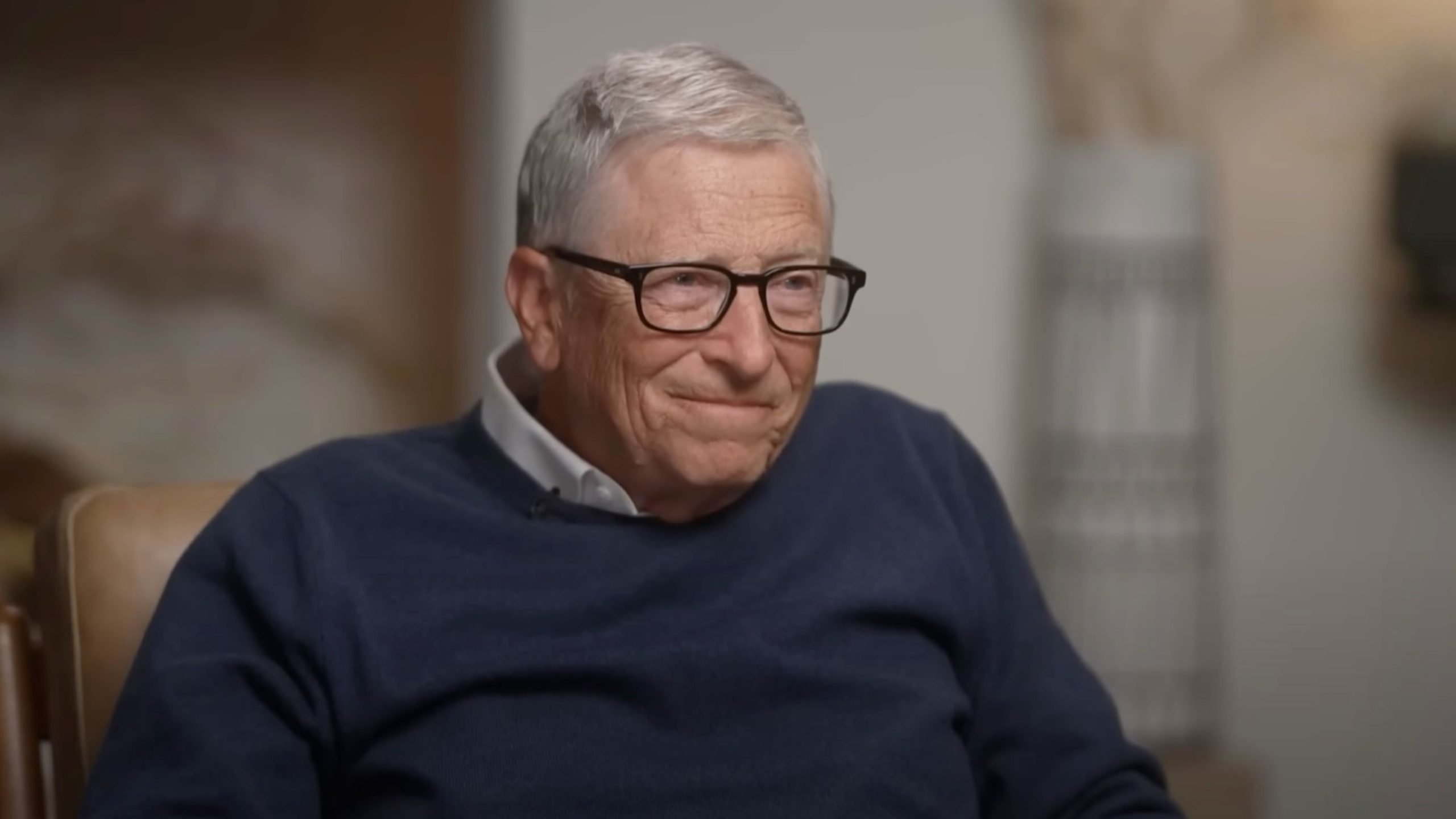 Bill Gates with gray hair and glasses wearing a navy blue sweater over a white collared shirt, sitting and smiling slightly indoors.
