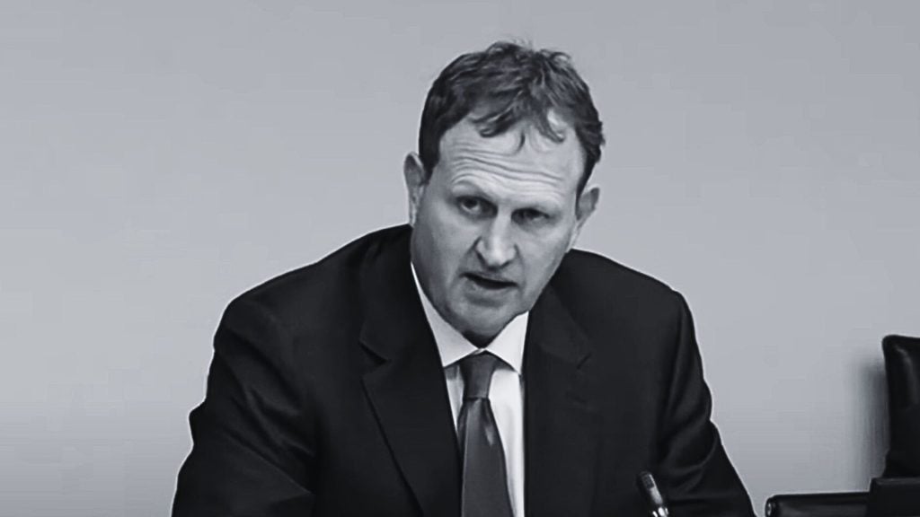 Ireland's Justice Minister Jim O'Callaghan in a suit and tie speaking, seated against a plain light-colored background in a formal setting.