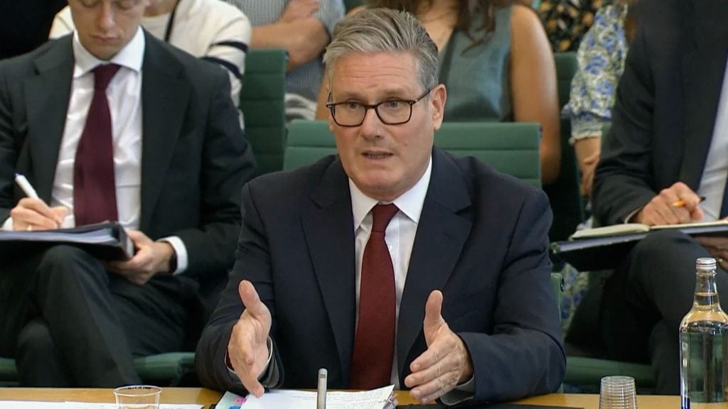 Starmer in a dark suit, white shirt, and maroon tie wearing glasses speaks gesturing with both hands at a table with papers and a microphone, with people sitting and taking notes in the background.