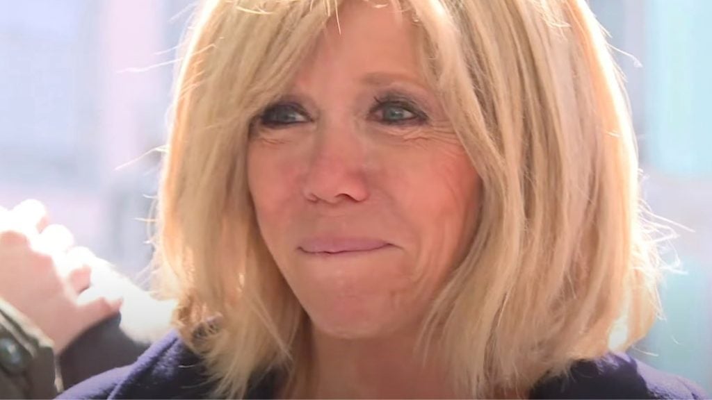 Close-up of a woman with shoulder-length blonde hair, slightly smiling with her lips pressed together and wearing dark eye makeup, with a light background and a hand resting on her shoulder