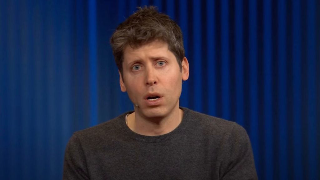 Sam Altman brown hair and blue eyes wearing a dark gray sweater speaking with a serious expression against a blue curtain background.