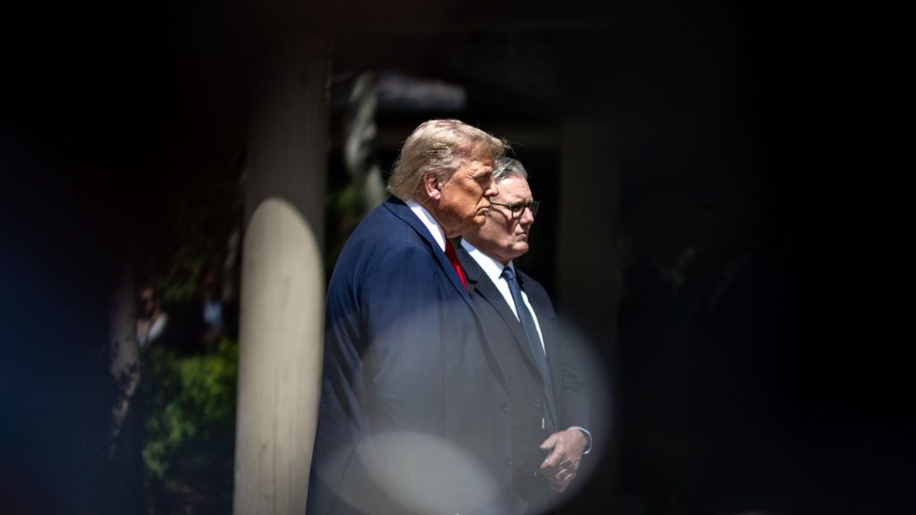 Starmer and Trump in suits standing side by side, one with light hair wearing a red tie and the other with glasses wearing a blue tie, with blurred dark foreground framing the image.