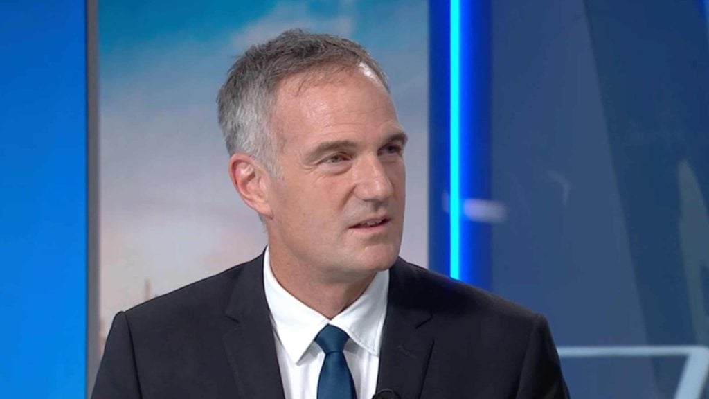 Peter Kyle with short gray hair wearing a dark suit, white shirt, and blue tie, looking slightly to the side in a studio setting with blue and gray background elements.