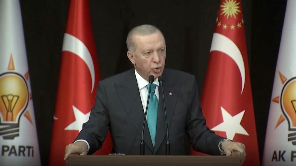 Erdogan in a suit with a teal tie speaking at a podium with two microphones, flanked by two Turkish flags and two flags with a lightbulb symbol and AK PARTI text.