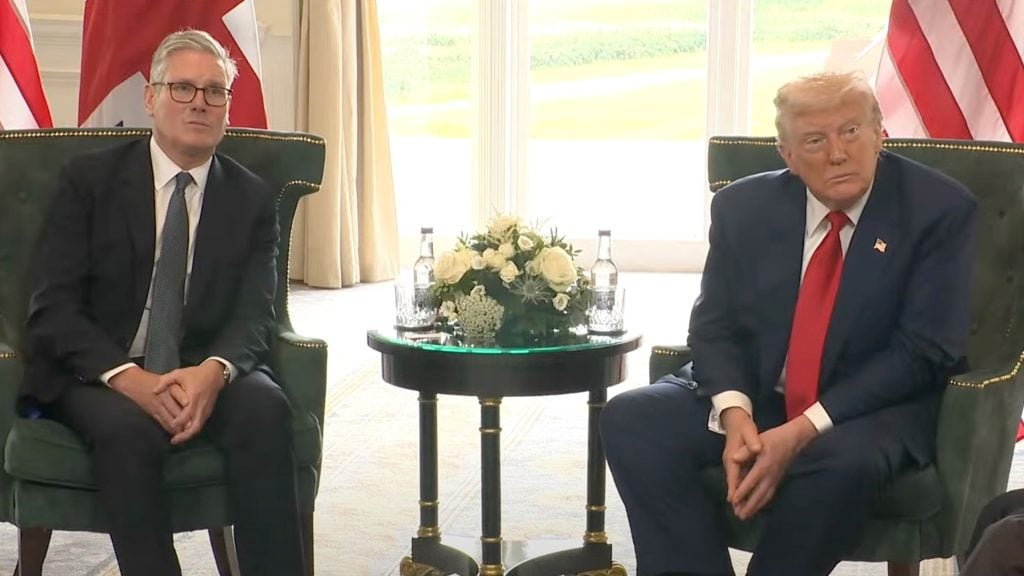 Starmer and Trump in suits sitting on green armchairs with a small round table with flowers between them, U.S. and Canada flags partially visible behind them.