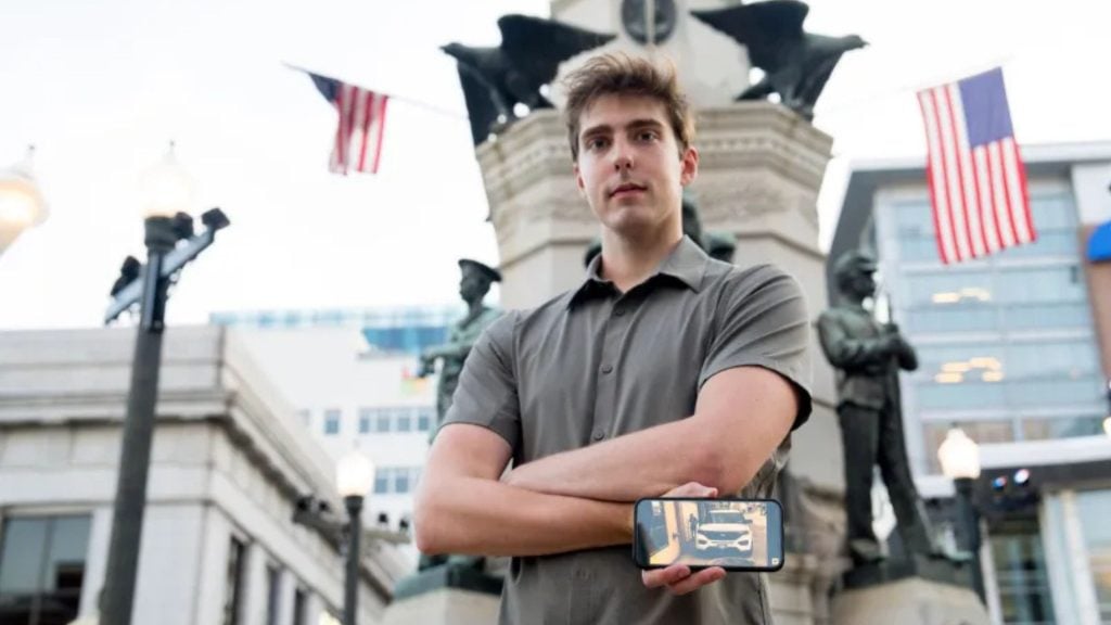 Phil Rishel stands outdoors in front of a monument with statues and American flags, holding a smartphone displaying an image of a white car on a street.