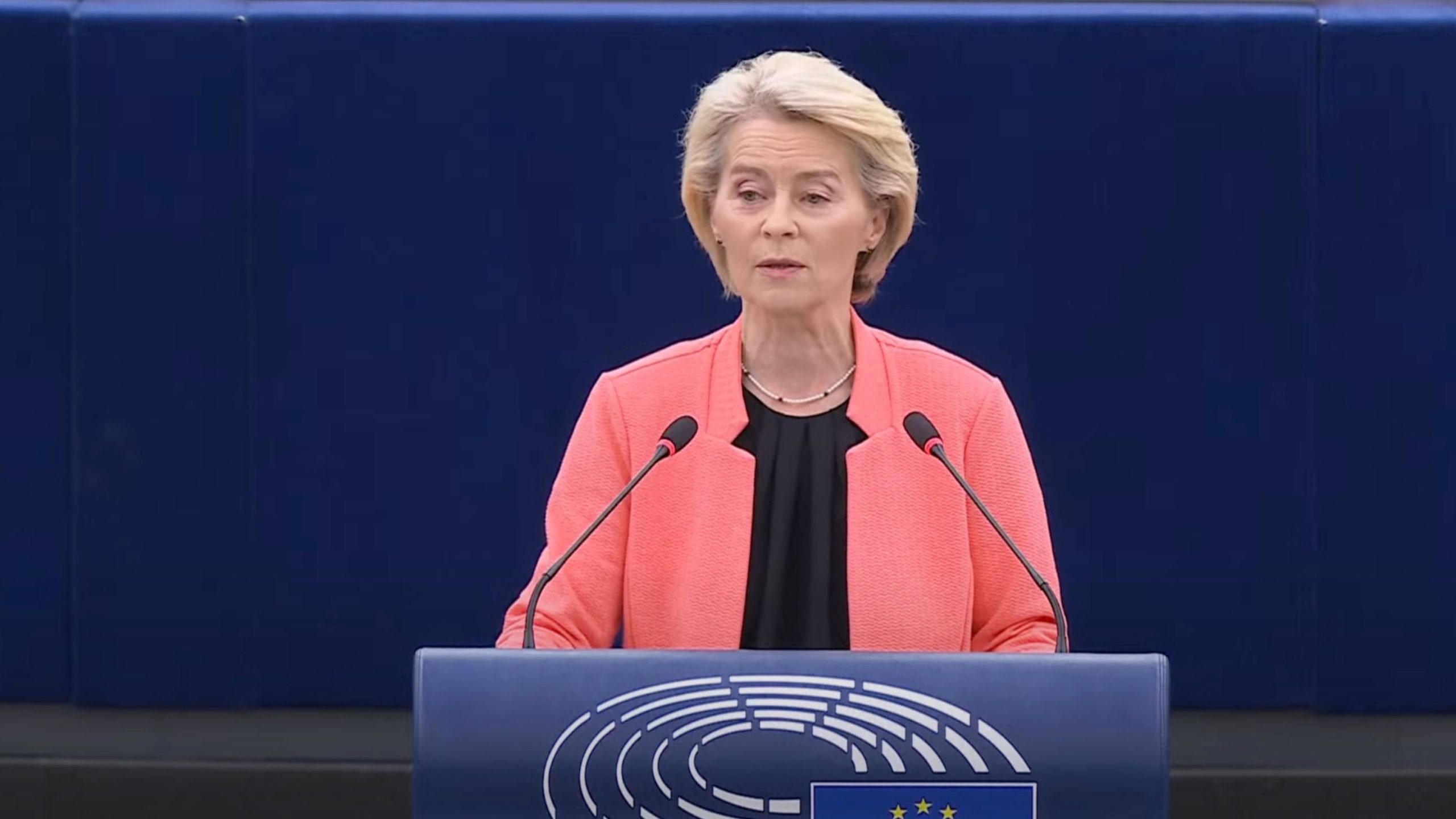 Ursula von der Leyen with short blonde hair wearing a coral jacket and black top speaking at a podium with two microphones, against a dark blue background with a symbol featuring white curved lines and a blue square with yellow stars.