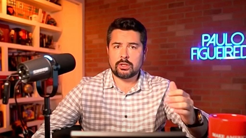 Figueiredo wearing a checkered shirt speaking into a microphone with a bookshelf and a neon sign that says PAULO FIGUEIREDO in the background.