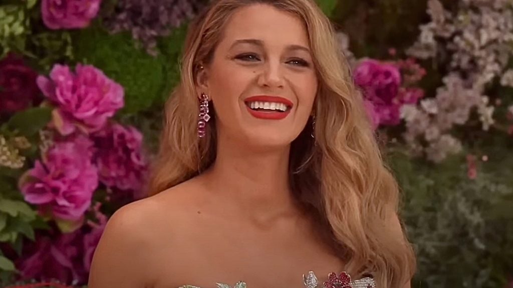 Blake Lively with long wavy blonde hair, wearing red lipstick and purple dangling earrings, dressed in a strapless outfit decorated with flowers, standing in front of a background filled with various pink and purple flowers.