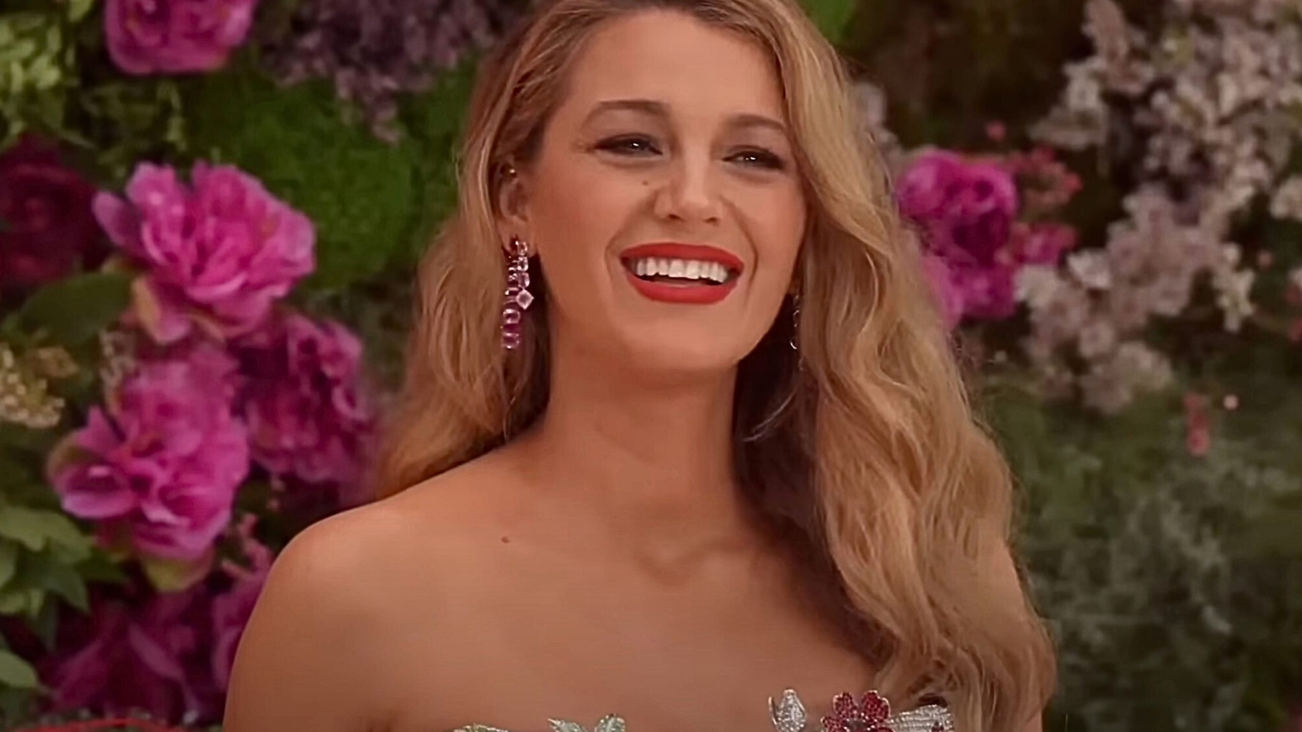 Blake Lively with long wavy blonde hair, wearing red lipstick and purple dangling earrings, dressed in a strapless outfit decorated with flowers, standing in front of a background filled with various pink and purple flowers.