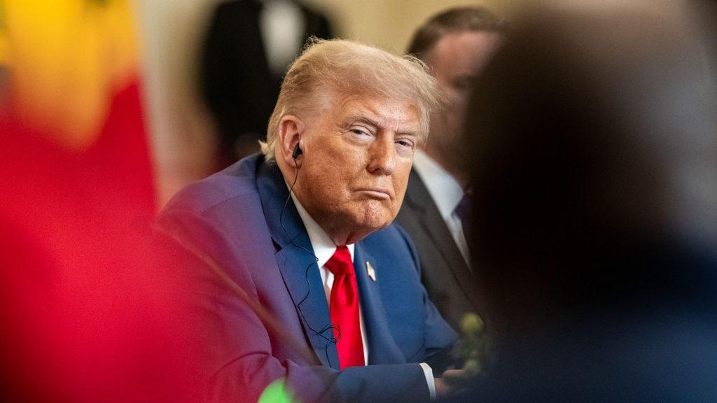 President Trump wearing a dark blue suit, white shirt, and red tie, looking directly at the camera with a serious expression and wearing a black earpiece, with blurred people and objects in the foreground and background.