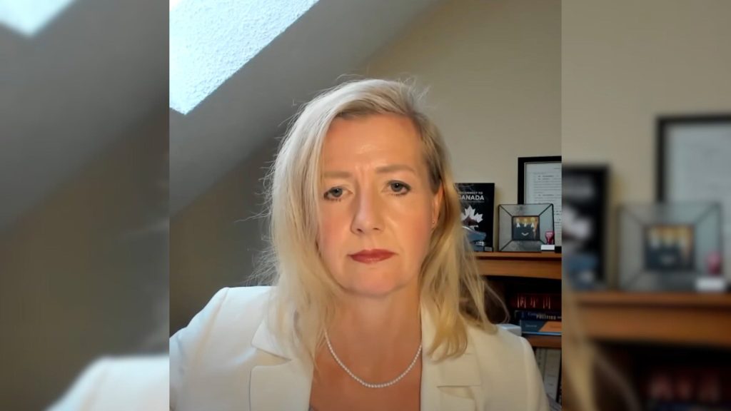 Eva Chipiuk with blonde hair wearing a white blazer and pearl necklace, sitting in front of a slanted ceiling with a bookshelf and framed pictures behind her.