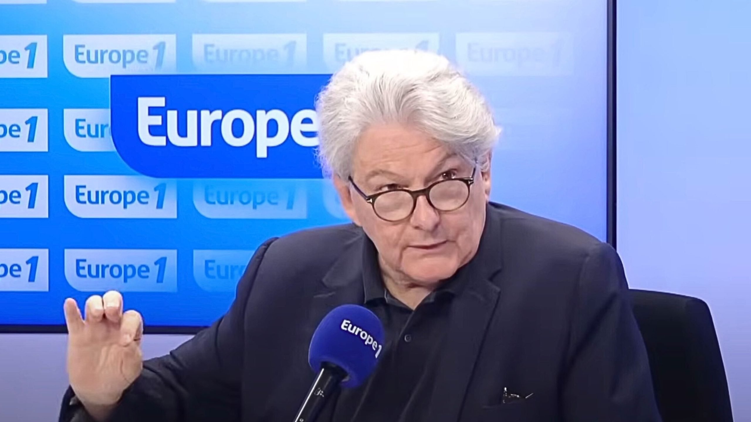 Breton with round glasses wearing a dark jacket, speaking into a blue Europe 1 microphone with his hand raised against a blue studio backdrop patterned with Europe 1 logos.