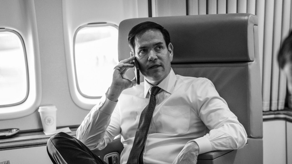 Rubio sitting in an airplane seat, speaking on a cellphone, with a cup and airplane windows visible in the background.