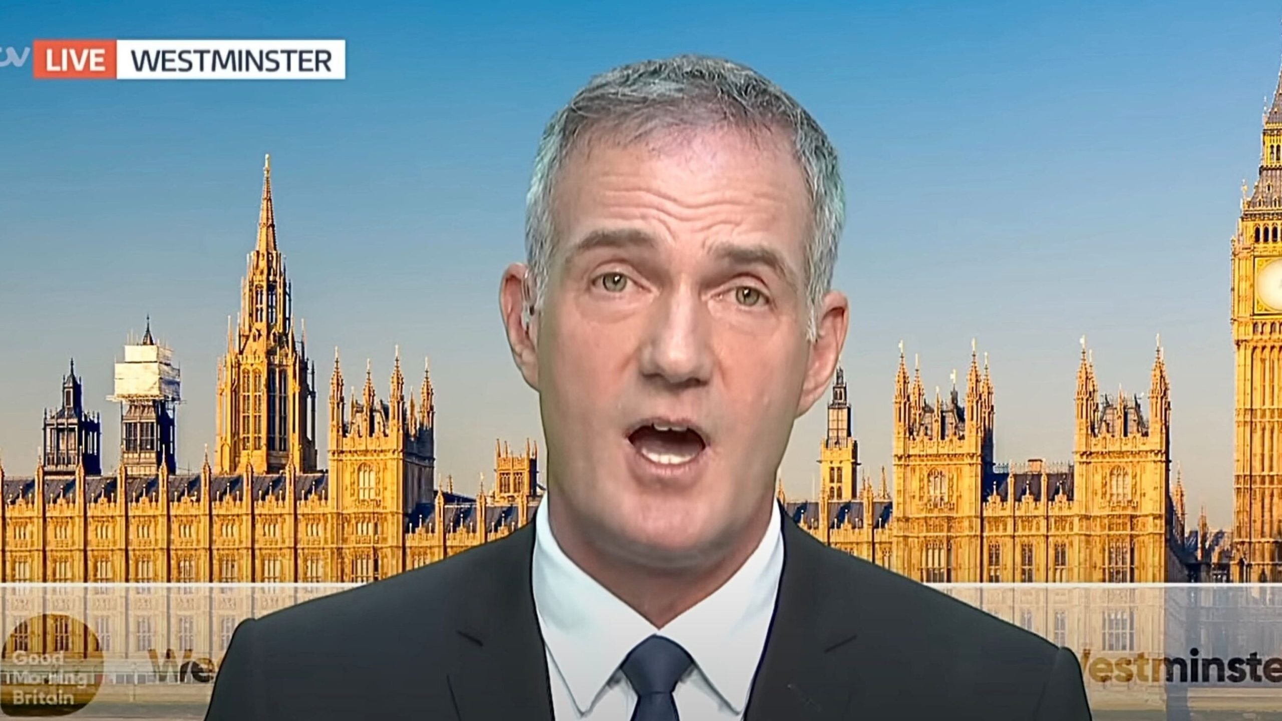 Kyle in a suit and tie speaking on a live ITV broadcast with the Houses of Parliament and Big Ben visible in the background at Westminster under a clear blue sky.