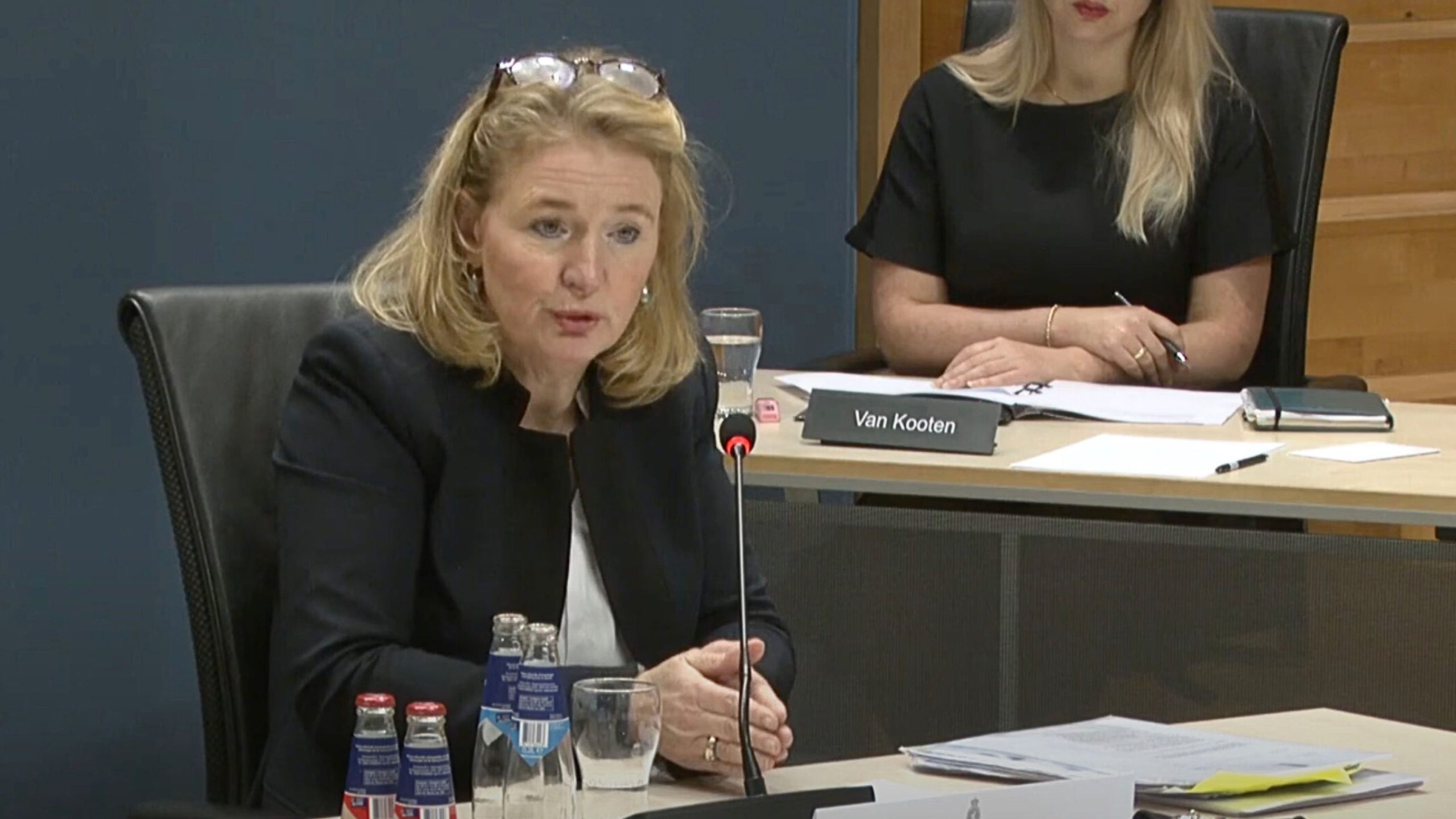 Manon Leijten in a dark blazer with glasses perched on her head speaking into a microphone at a conference table, with bottled drinks, a glass of water, stacks of papers and another woman seated behind her.