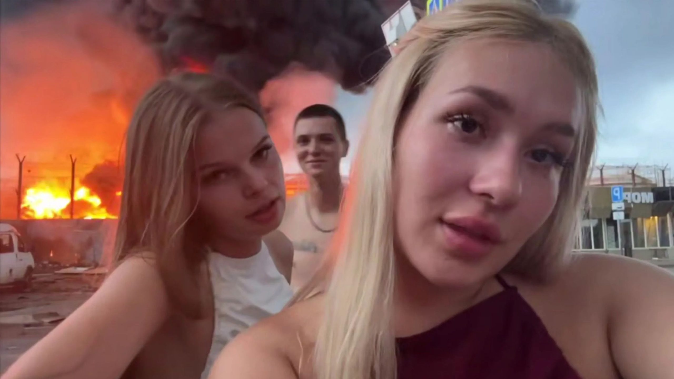Three young people taking a selfie outdoors with a large fire and thick black smoke rising in the background near a fence and some buildings.