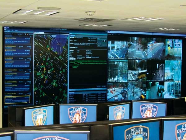 Control room video wall showing a city map, multiple live surveillance camera feeds, and rows of monitors displaying Police Department shields.