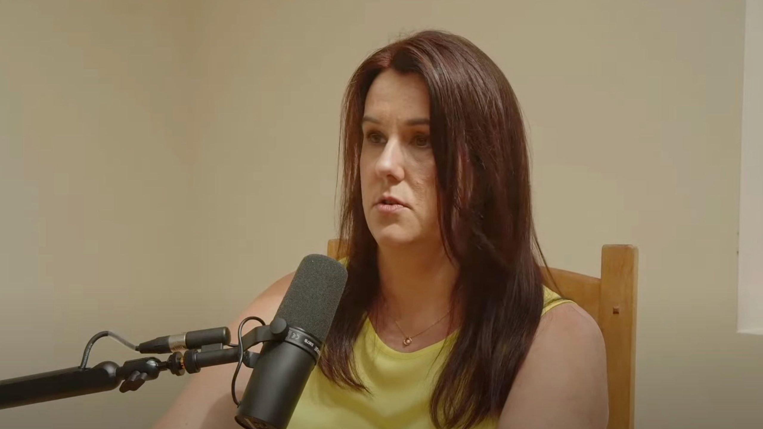 Lucy with dark brown hair wearing a yellow sleeveless top and a small pendant necklace, seated in a wooden chair and speaking into a black microphone on a boom arm against a plain beige wall with a serious expression.