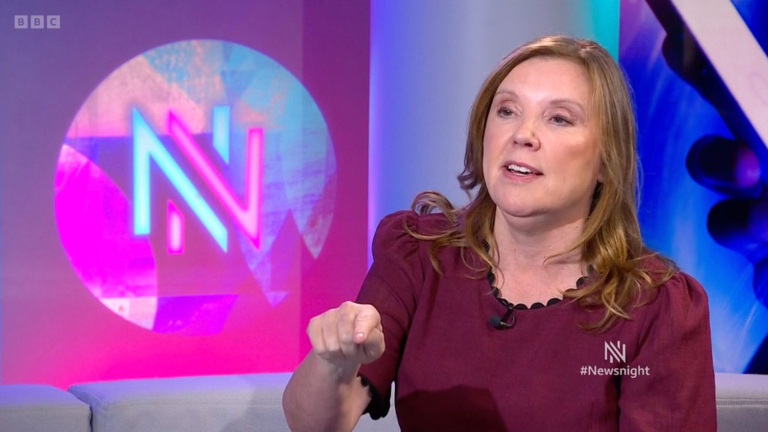 De Souza in a maroon top seated on a studio sofa, pointing toward the camera with a colorful neon circular 'N' logo and #Newsnight caption visible in the background.