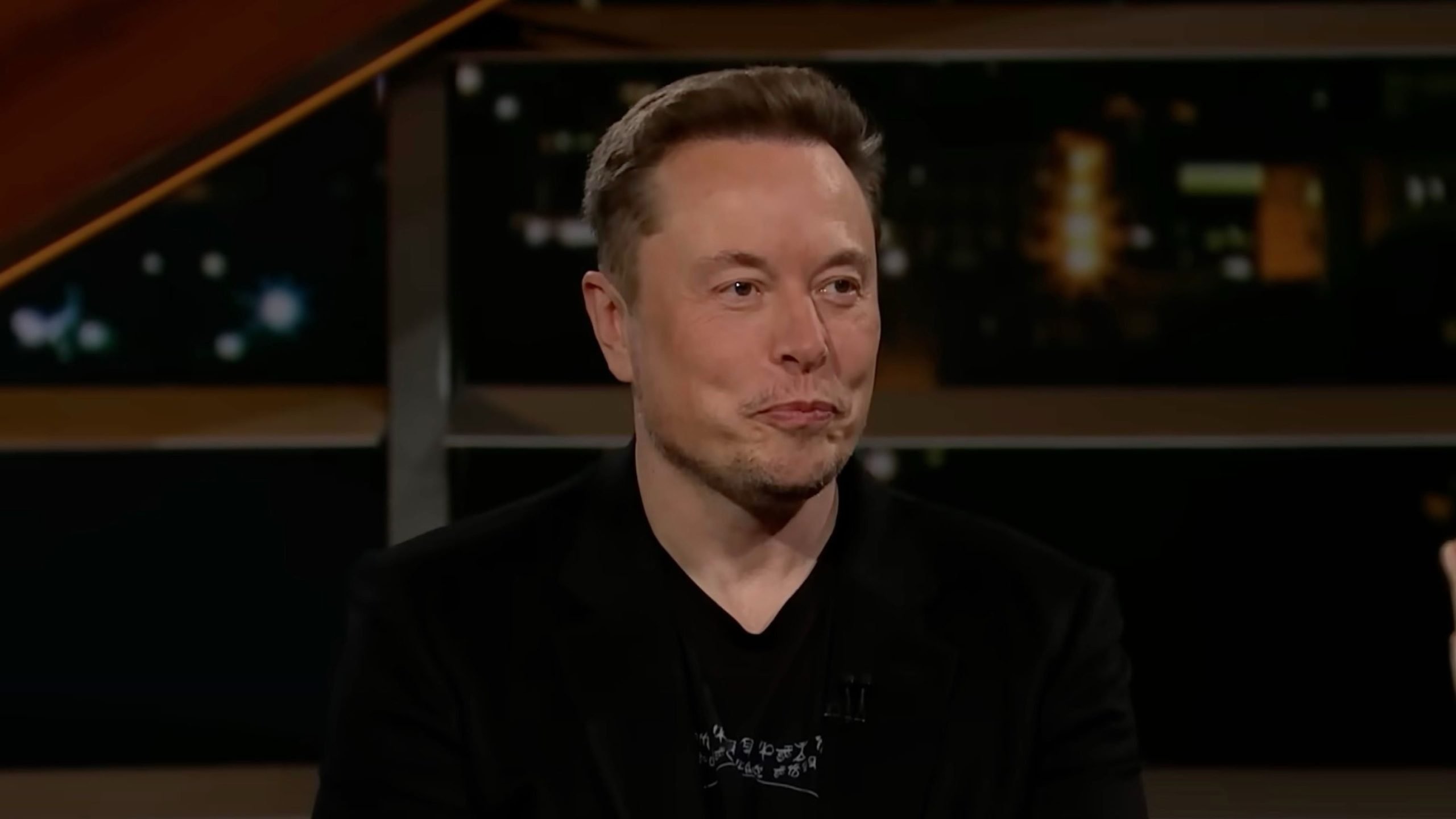 Elon Musk with short brown hair and light stubble wearing a black shirt and blazer, seated in a studio with a blurred background of lights and railings, lips pursed in a thoughtful expression.