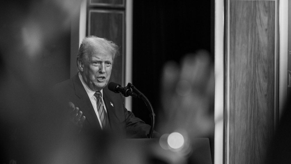 Black and white photo of Trump in a suit speaking into a microphone with a blurred hand in the foreground and wood-paneled wall in the background.