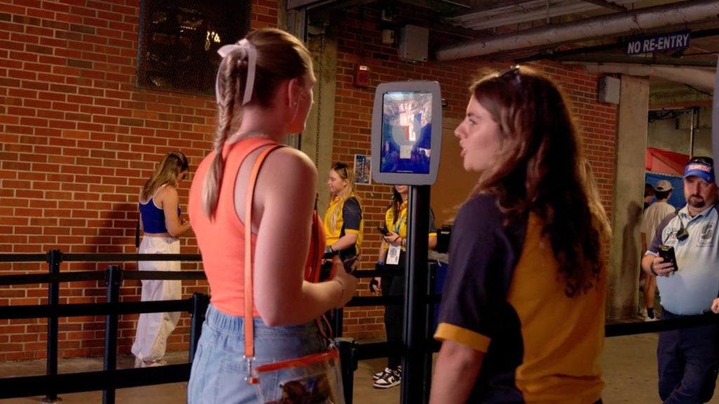 Two young women stand in a brick-walled stadium corridor at a kiosk — one with a braided ribbon in an orange tank top and clear crossbody bag faces the screen while a staff member in a yellow-and-black uniform talks, with other staff and a security officer nearby under a "No Re-Entry" sign.