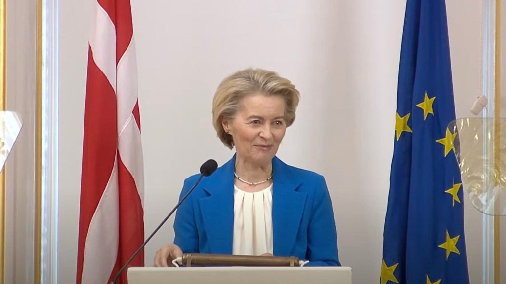 Von De Leyen with short blonde hair in a blue blazer speaking at a podium with a microphone, flanked by a red-and-white vertical flag and a blue flag decorated with yellow stars.
