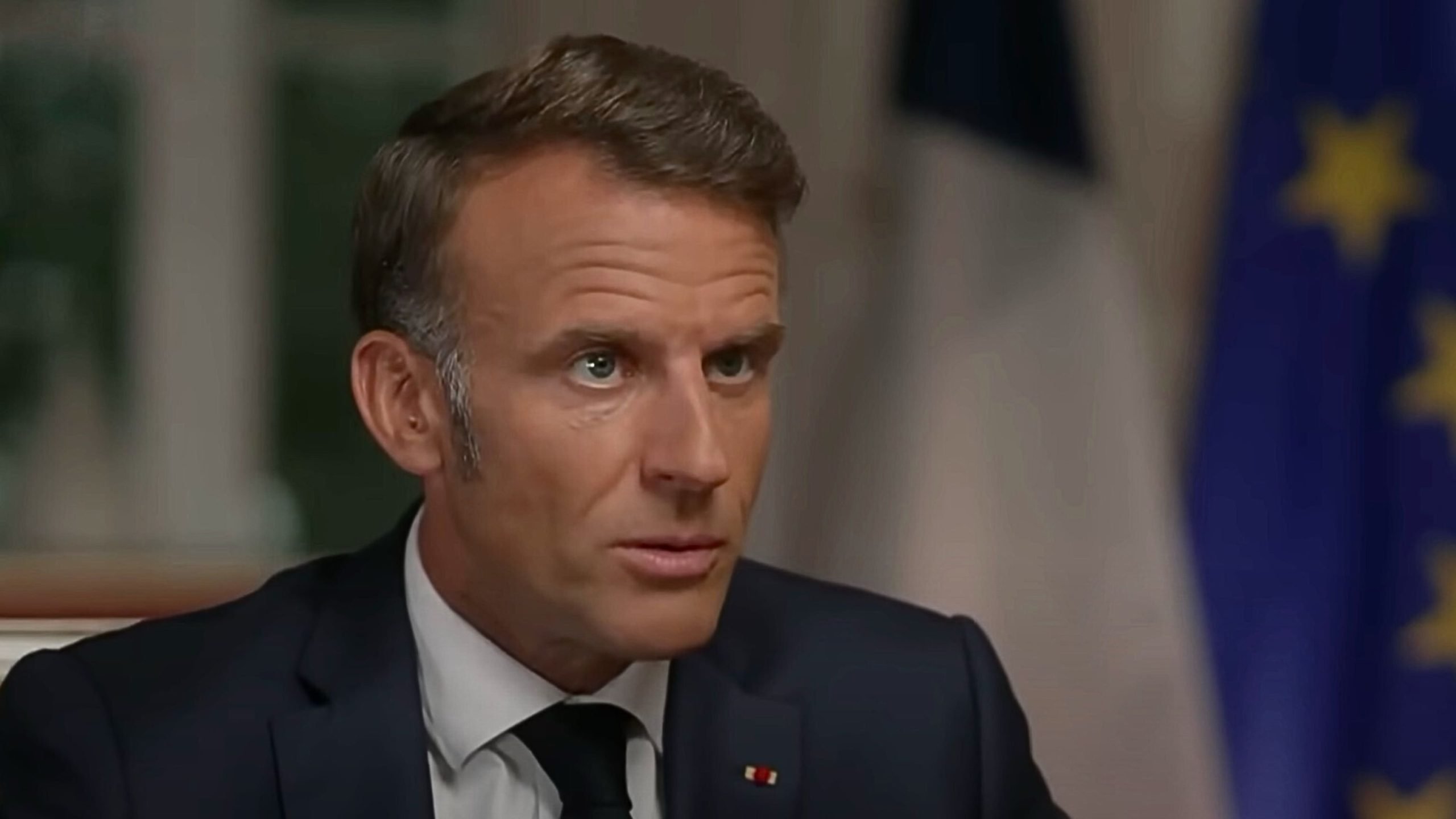 Macron with short brown hair wearing a dark suit, white shirt and black tie, seated indoors with a small tricolored lapel pin and blurred national and European Union flags in the background.