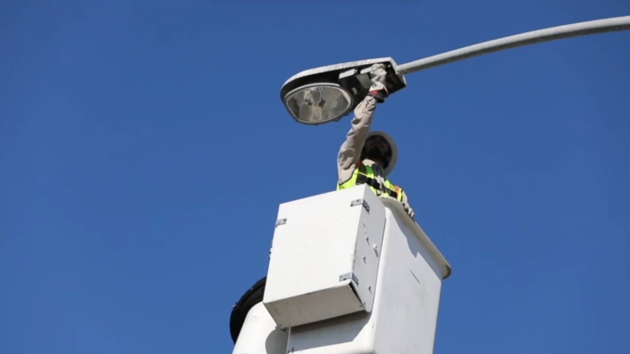Utility worker in a hard hat and reflective vest stands in a white bucket lift reaching up to repair a streetlight against a clear blue sky.