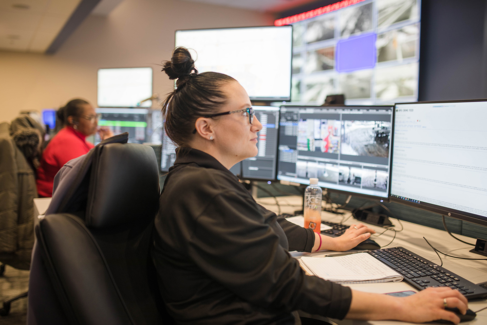 Woman wearing glasses and her hair in a bun sits at a control-center workstation monitoring multiple large computer screens showing surveillance footage and data, with a notebook and bottled drink on the desk and another operator in the background.