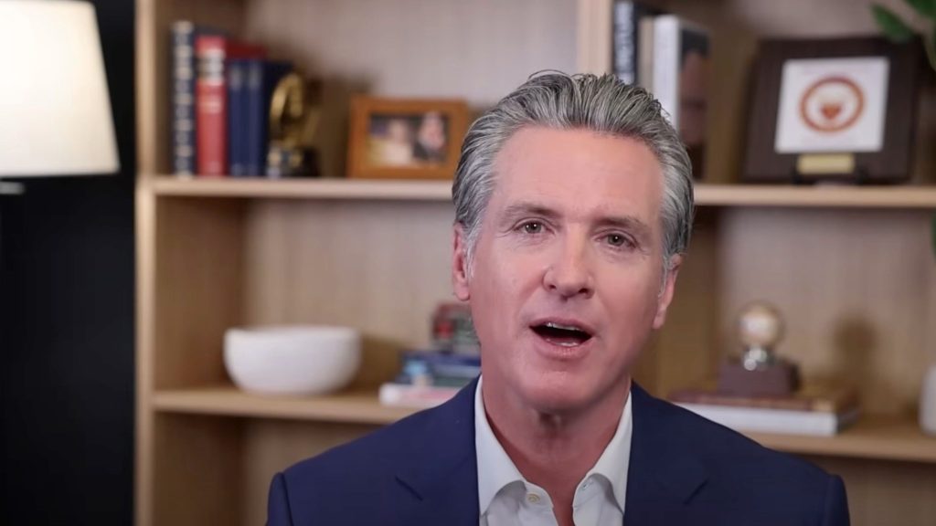 Newsom wearing a dark blue suit and white shirt speaking in front of a bookshelf with books, a lamp, and framed items.
