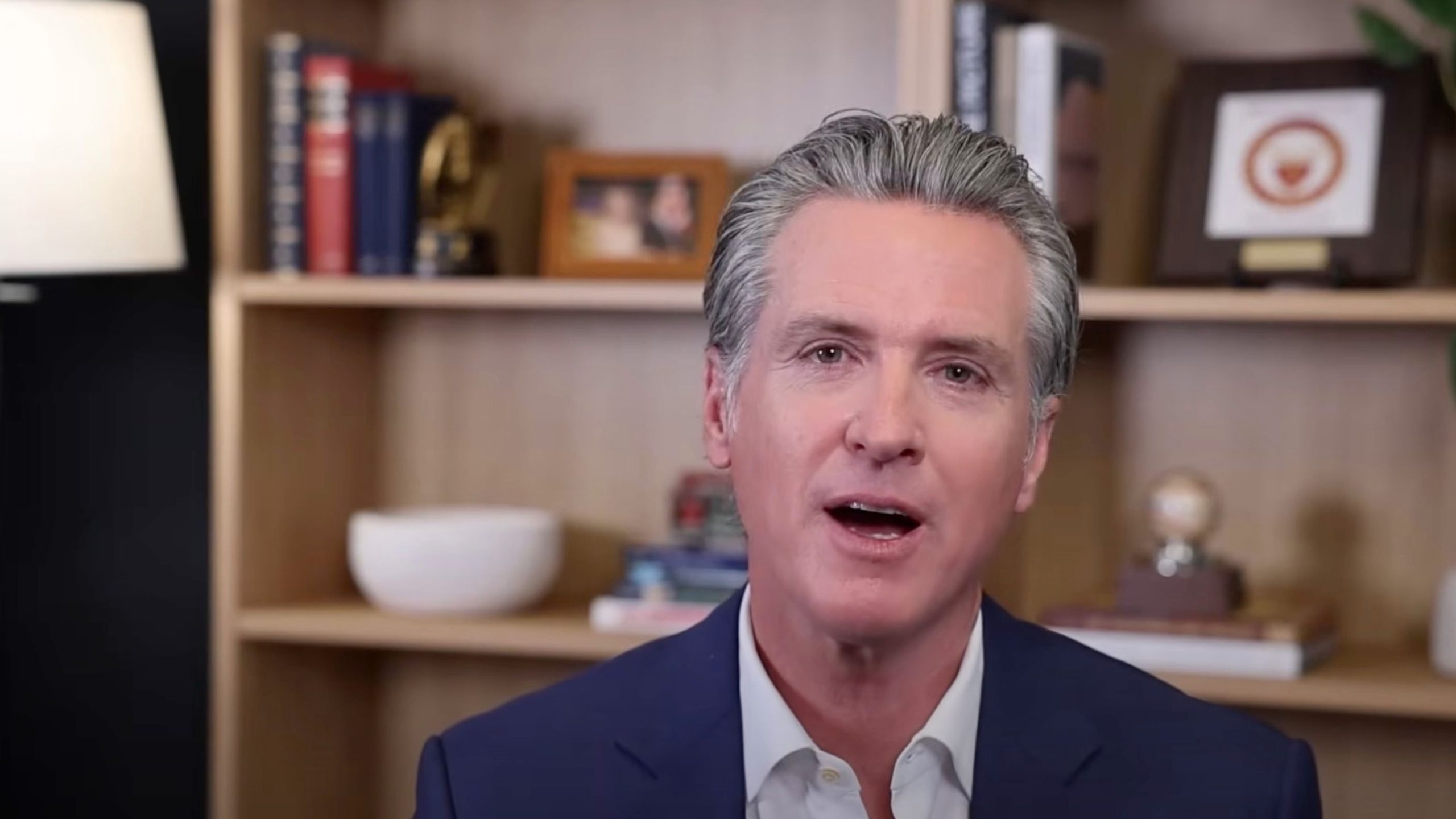 Newsom wearing a dark blue suit and white shirt speaking in front of a bookshelf with books, a lamp, and framed items.